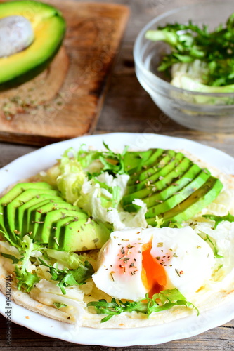 Poached egg, avocado slices, salad mix, sauce, spices in a flour tortilla. Avocado half on a wooden board, salad mix in a glass bowl on a table. Healthy and diet breakfast idea. Vertical photo