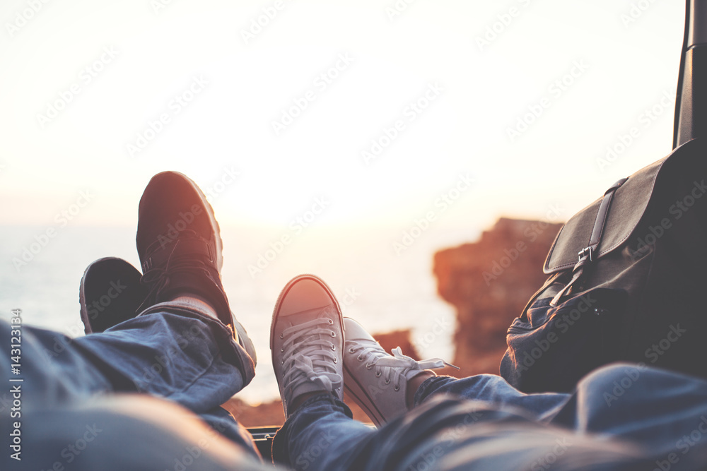 Couple lying at sunset near the ocean in the van's trunk, close-up of shoes of woman and man, romantic relationships - obrazy, fototapety, plakaty 