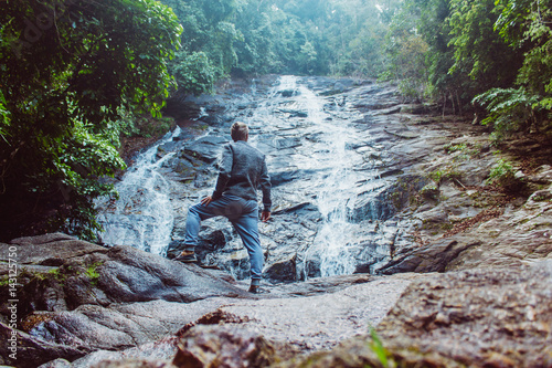 Male posing against waterfall . Back view of young man standing on rock near beautiful water stream flowing down stiff slope.