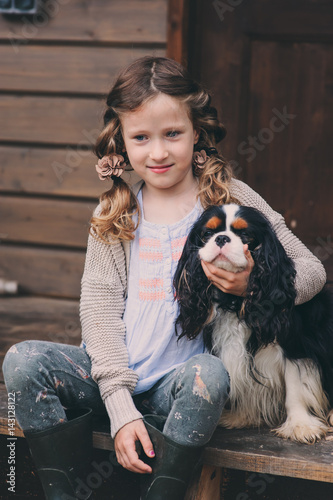 kid girl playing with her spaniel dog, sitting on stairs at wooden log cabin