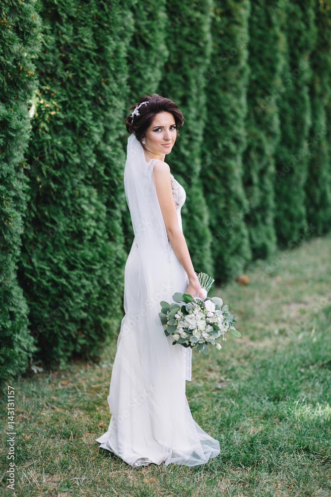 Brunette bride looks over her shoulder standing before green trees in the park