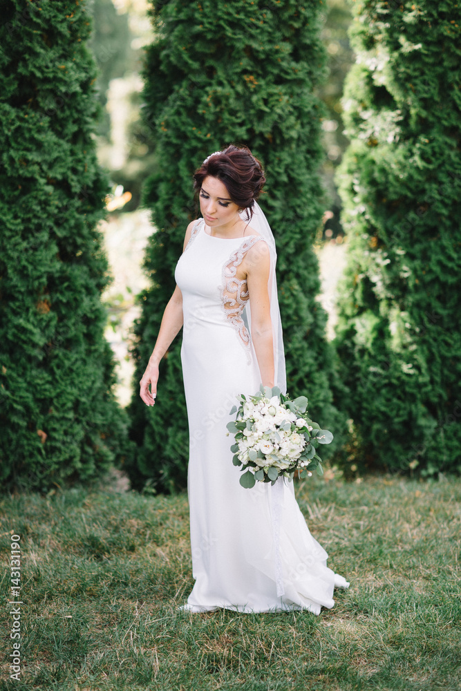 Bride in elegant gown holds hand with bouquet down standing on the lawn Bride in elegant gown holds hand with bouquet down standing on the lawn