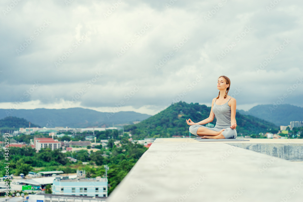 Yoga on rooftop. Happy young woman in lotus pose on roof with city and mountains view.