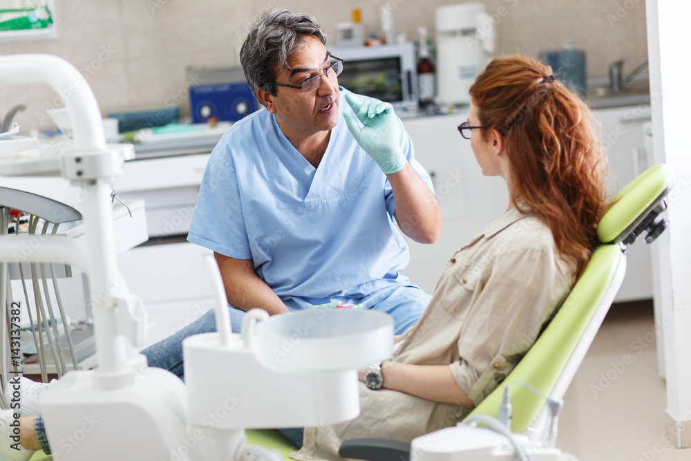 A professional dentist, equipped with a bright smile, converses with his red-haired female patient, carefully explaining the upcoming treatment, and ensuring her comfort throughout the process. A professional dentist, equipped with a bright smile, converses with his red-haired female patient, carefully explaining the upcoming treatment, and ensuring her comfort throughout the process.