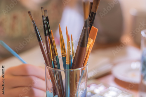 Paint brushes in glass on the table in a workshop. Master drawing at background.Close up.