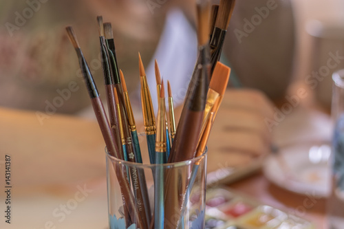 Paint brushes in glass on the table in a workshop. Master drawing at background.Close up.