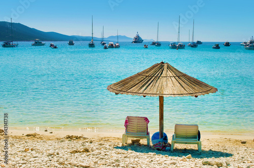 One yellow straw umbrella and two chairs on the pebble sandy beach. Many yachts in the azure blue sea lagoon in a bay near the coast of an island. Blue cloudless sky. Dugi otok, Croatia