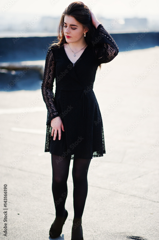 Portrait brunette girl with red lips wearing a black dress, tights and  shoes on heels posed on the roof. Street fashion model. Stock Photo