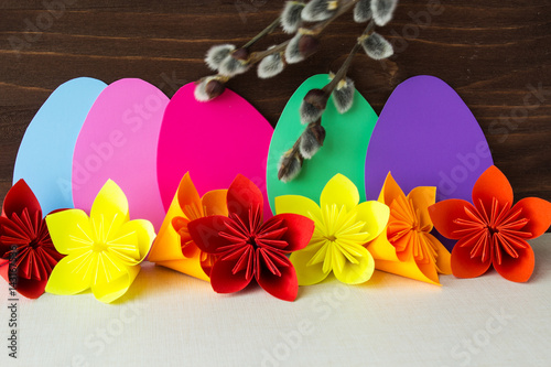 Decorative composition of Easter eggs, paper flowers and willow branches on a wooden background. Selective focus.