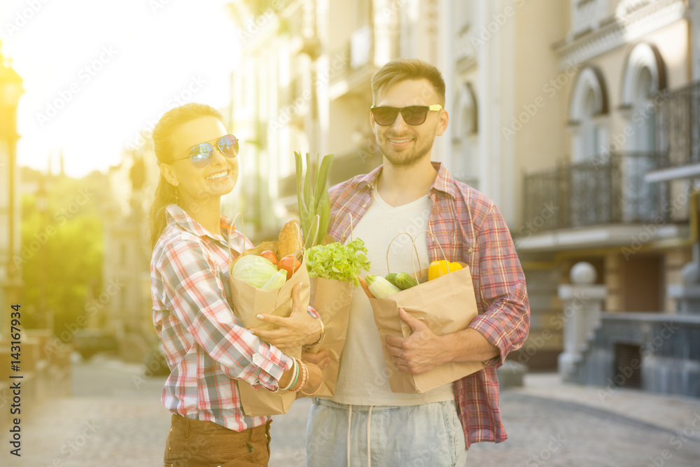 Fototapeta premium Happy couple after shopping. Man and woman in sunglasses carrying bags full of different food. Toned image.