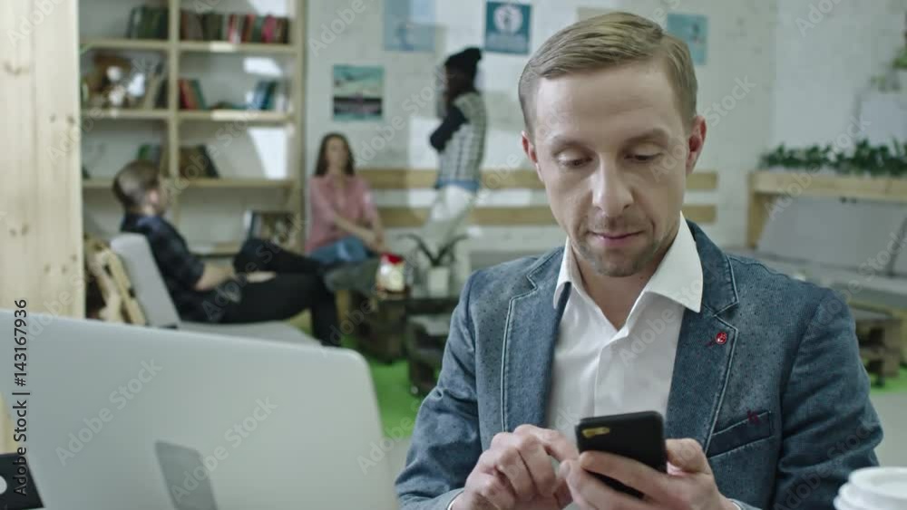 Tilt up of hipster businessman typing on laptop at desk and then answering phone call; young colleagues chatting in background Tilt up of hipster businessman typing on laptop at desk and then answering phone call; young colleagues chatting in background