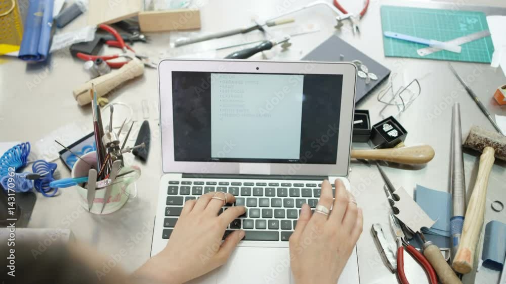  Jewelry designer at workbench making a to do list on computer. Close up on hands, no faces. 