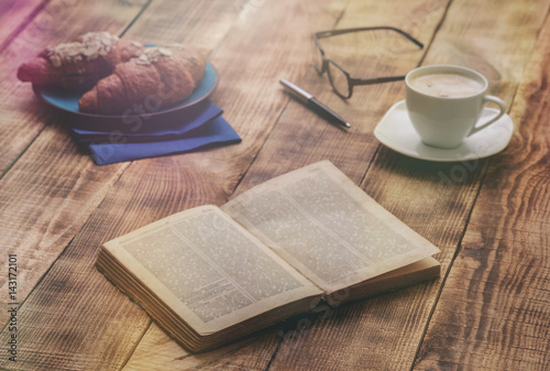 Open old book on wooden table with croissants and coffee