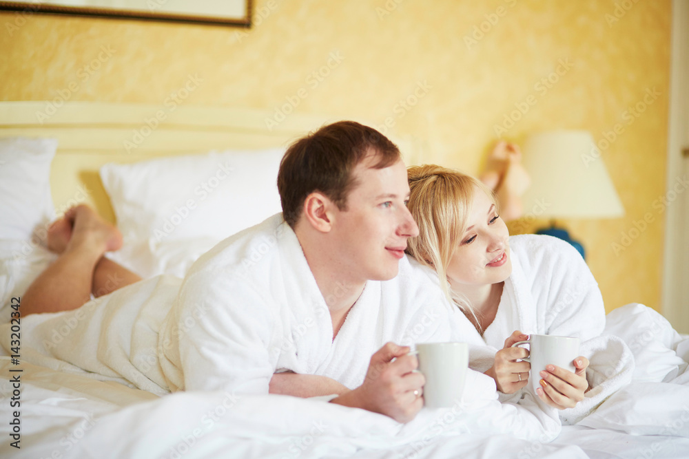 Fototapeta premium Couple in white bathrobes drinking coffee together in bed