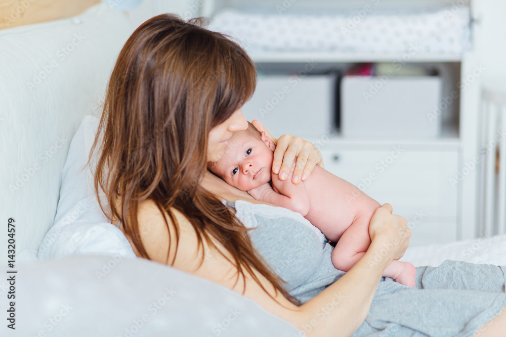 Mother holding newborn baby and lying on the bed in cozy room. Side view. Child Parenting Love Concept - obrazy, fototapety, plakaty 