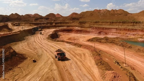 Two dump trucks move in the quarry. Excavators. Ore extraction. Mining. Bouxite. Panorama. Red earth. Azure artificial lake. View from the drone.