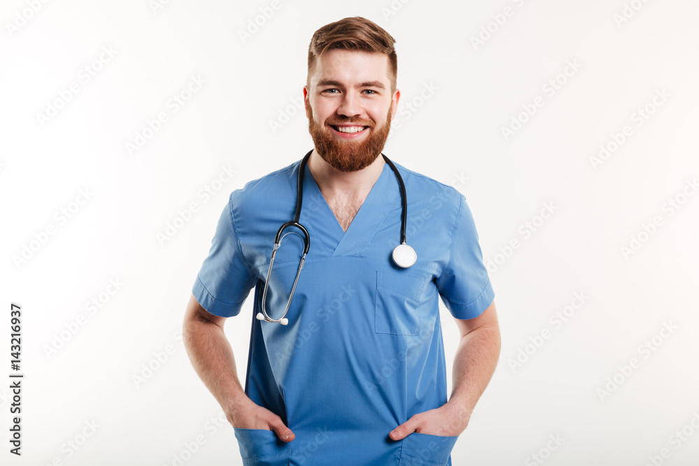 Smiling male doctor standing with stethoscope and holding hands in pockets Smiling male doctor standing with stethoscope and holding hands in pockets