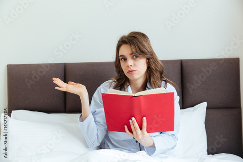 Confused young woman wearing pajamas and holding book in bed © Drobot Dean