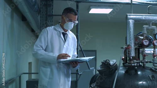 Side view video of male scientist in white lab coat, goggles and mask standing next to productional equipment checking indicators and writing in journal photo