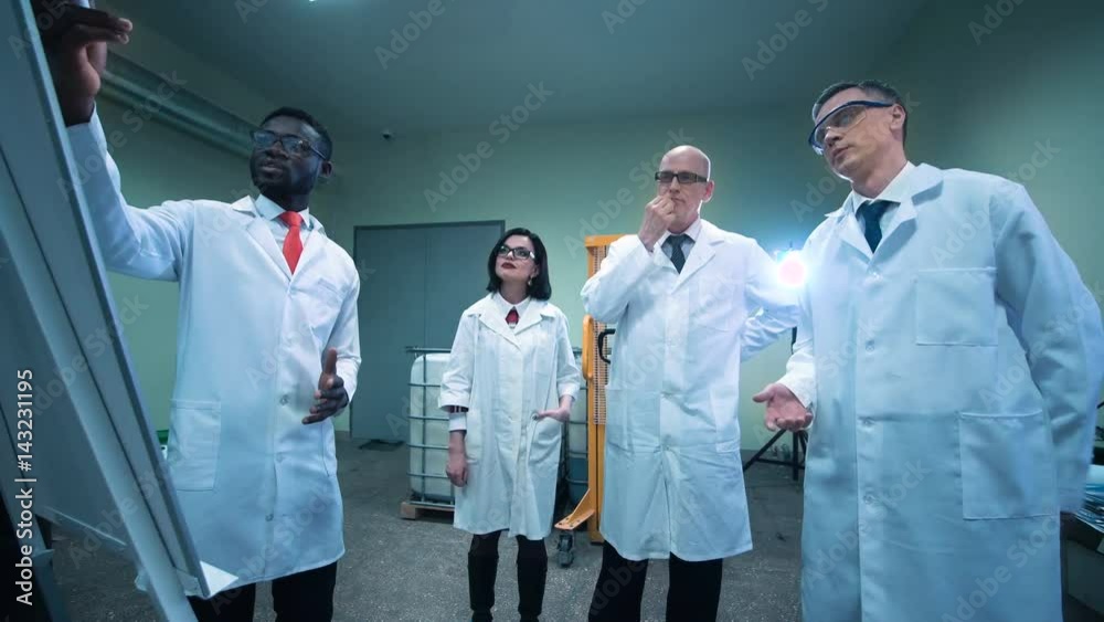 Four scientists in white lab coats standing indoors and discussing project that black man showing on white board