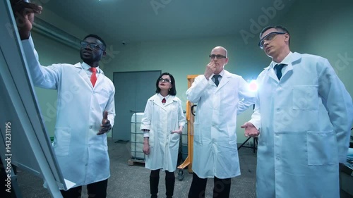 Four scientists in white lab coats standing indoors and discussing project that black man showing on white board photo