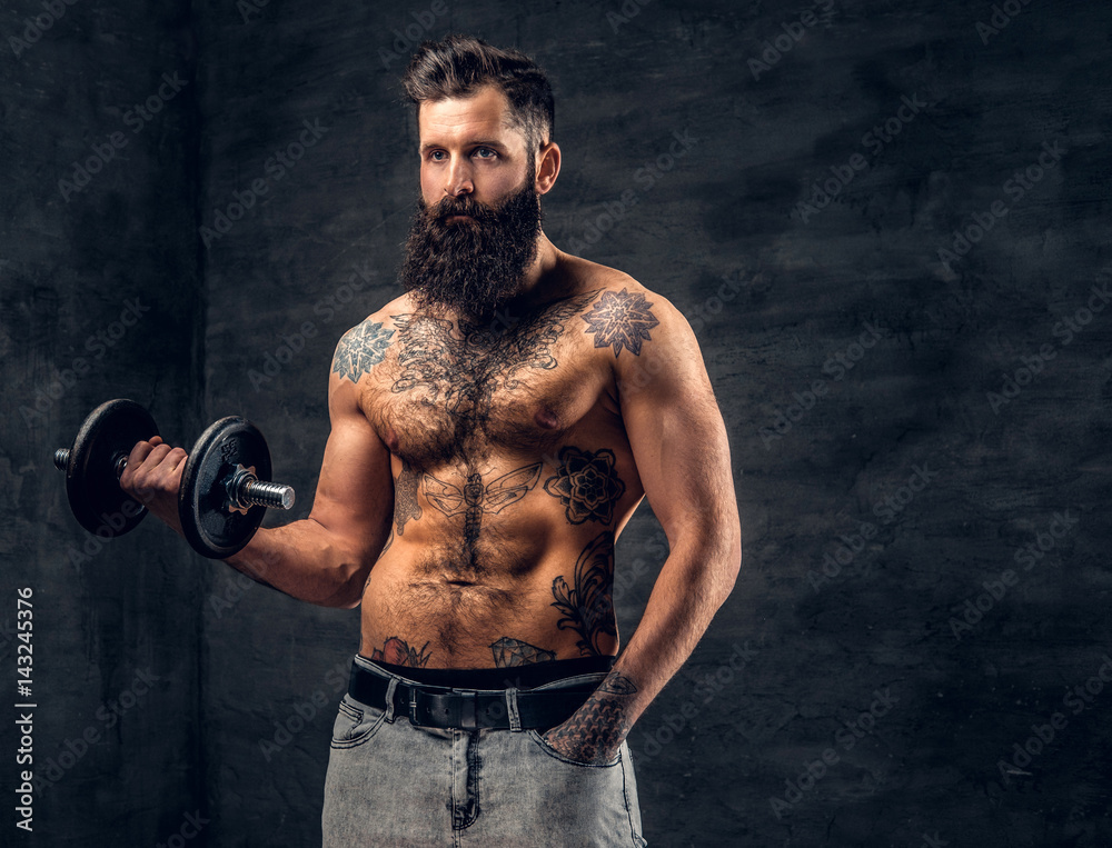 Shirtless bearded male with tattooed torso doing workout  with dumbbell.