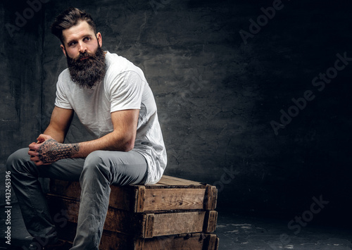 Bearded male with tattoo on arm dressed in a white t shirt sits on a wooden box.