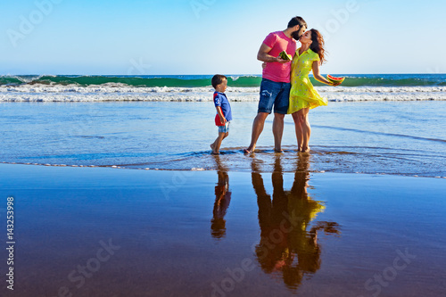 Happy family - father, mother, baby son walk by water pool on black sand beach with sea surf. People have picnic - eat tropical fruits. Travel, active lifestyle, parents with child on summer vacation