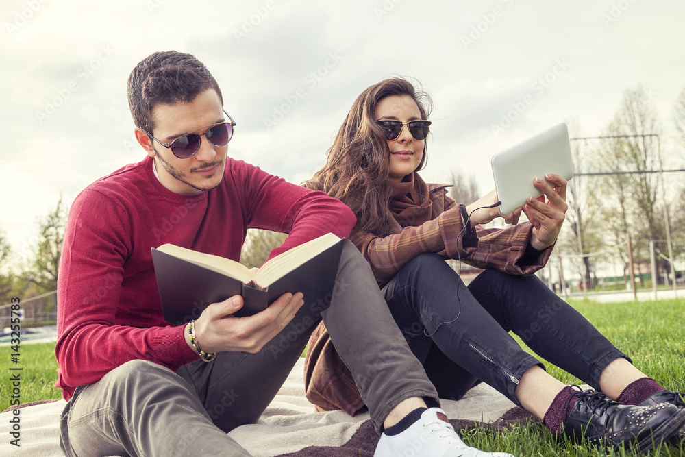 nice couple boyfriends relaxing on meadow looking at book and smart phone