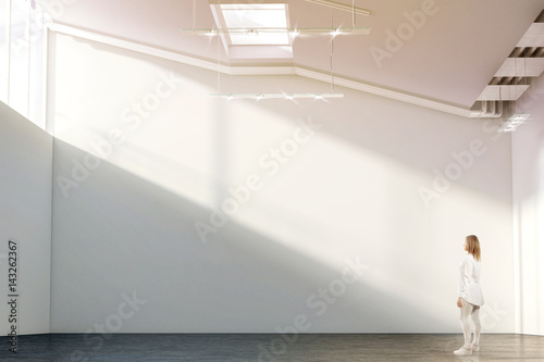 Woman walking near blank white wall mockup in modern gallery with sunrise. Girl admires a clear big stand mock up in museum with contemporary art exhibitions. Large hall interior, sunset from windows photo