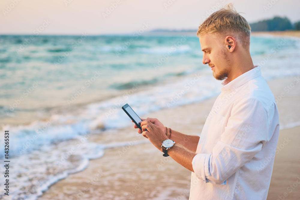 Vacation and technology. Portrait of young handsome man using smartphone on the sea shore.