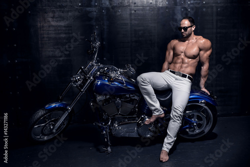 Sexy rich handsome attractive sporty muscular shirtless bodybuilder and fitness model with perfect torso body in white jeans sits on a beautiful blue shiny luxury motorcycle