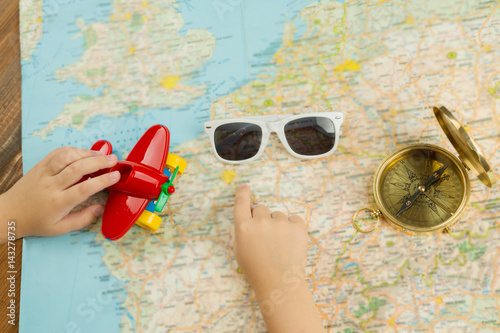 Dreaming about summer holiday trip. Kid hand pointing to the map. Sun glasses, compass and toy red plane. Top view 