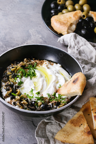 labneh middle eastern lebanese cream cheese dip with olive oil, salt, herbs, olives tapenade served in black bowl with traditional pita bread over gray texture metal background.