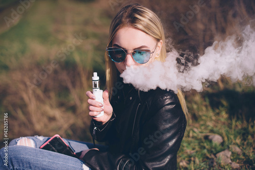 blonde girl vaping outdoors . female model smoking fruit flavored e-liquid or e-juice with vaporizer device or e-cig.Modern gadget for smokers photo