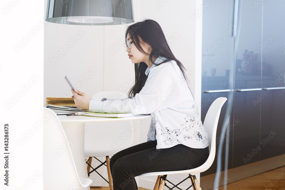 View from the side photo of young beautiful asian woman with long dark hair wearing glasses and white shirt and surfing the web by a digital tablet. Student girl is communicating with friends. View from the side photo of young beautiful asian woman with long dark hair wearing glasses and white shirt and surfing the web by a digital tablet. Student girl is communicating with friends.