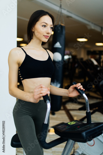 Young fit woman using an elliptic trainer in a fitness center and smiling. Portrait of fitness girl in the gym near a window, lifestyle concept.