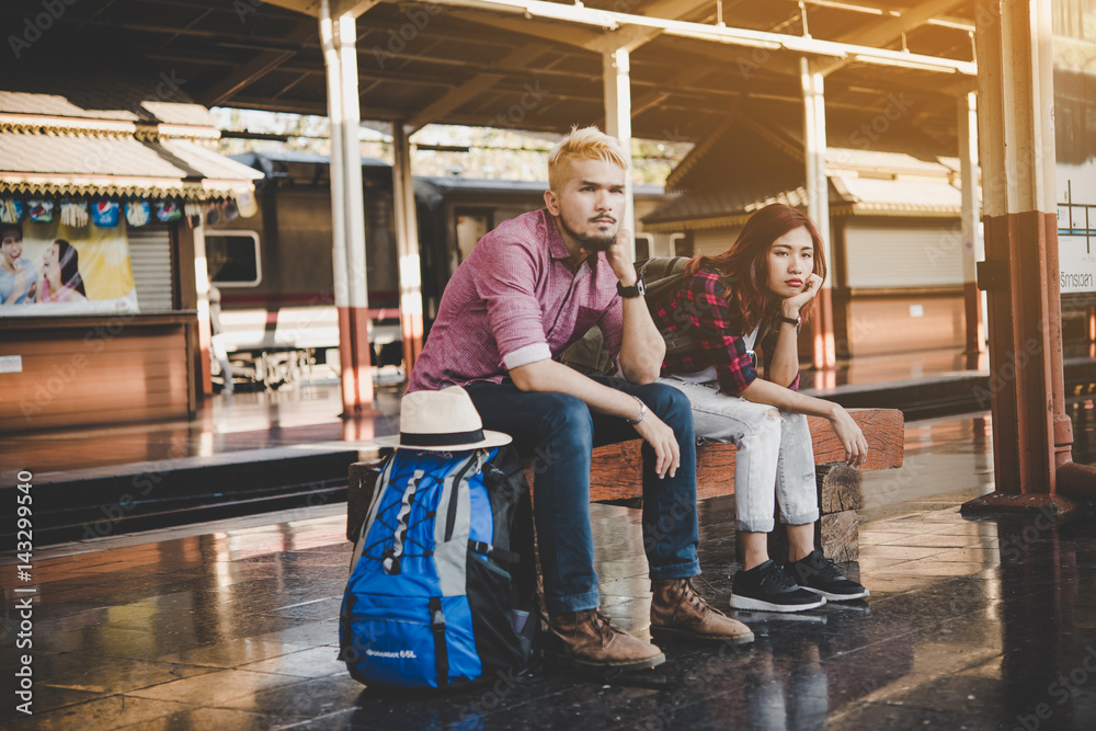 Hipster couple sitting on bench at the train station. Two young tourist are waiting to get on the train and begin their journey. Travel concept.