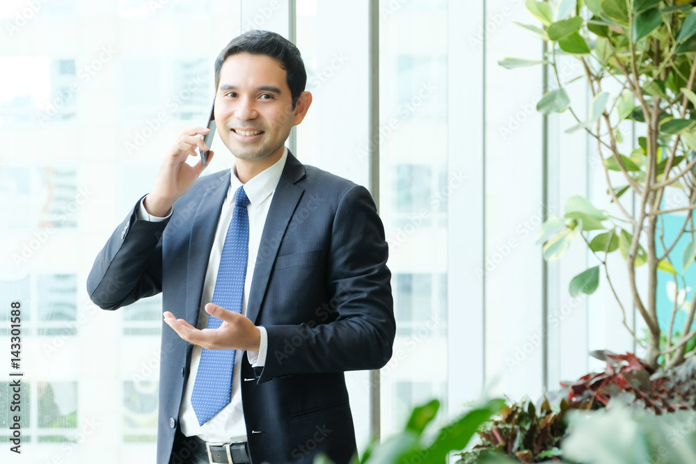 Business man talking smart phone with happy smiling face standing by windows, inside eco office building background, businessman on smart phone