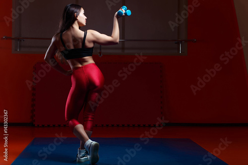 Young muscular woman bodybuilder with dumbbell in hand demonstrating body in gym, view from the back