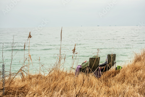 Folding chairs and a couple sitting on the seashore. Calm sea view. The shore is covered with dry grass. Early spring. 