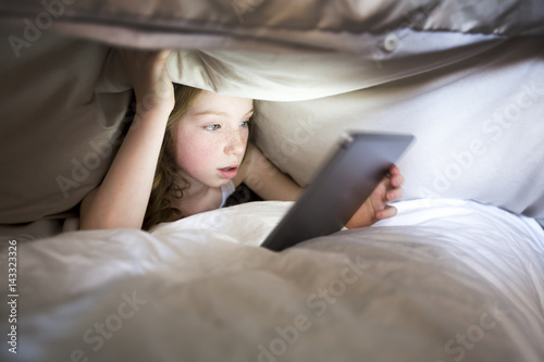 Cute little girl alone with tablet computer under blanket at night in a dark room