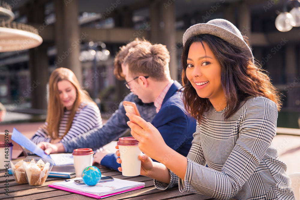 Fototapeta premium Happy students sit in time of a break and discuss about their tasks, with coffee, with a phones, with the folders, with the foods and one of them keeps a phone and cup of coffee in her hands, outdoors
