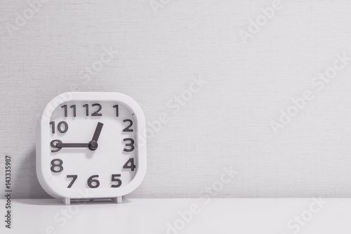 Closeup white clock for decorate show a quarter to one o'clock or 12:45 p.m. on white wood desk and cream wallpaper textured background in black and white tone with copy space
