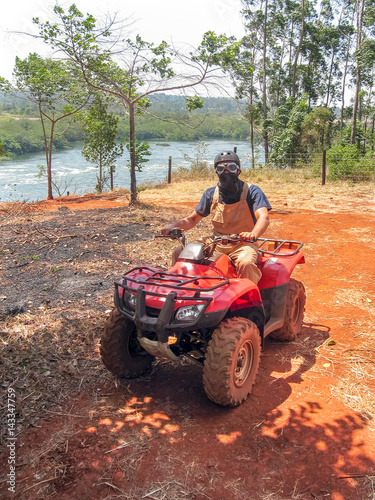 Man with kerchief on face sits on quad bike against Victoria Nile River background. Jinja, Uganda, Eastern Africa. 