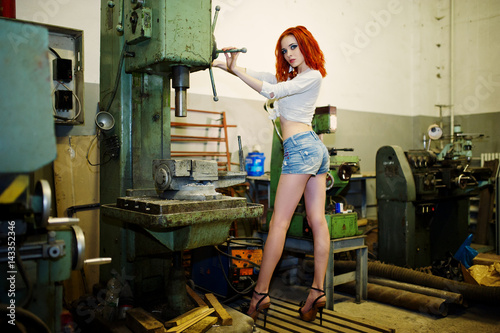Red haired girl wear on short denim shorts and white blouse posed at industrial machine at the factory. photo