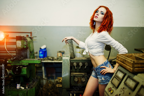Red haired girl wear on short denim shorts and white blouse with steel spring at hand posed at industrial machine at the factory. photo