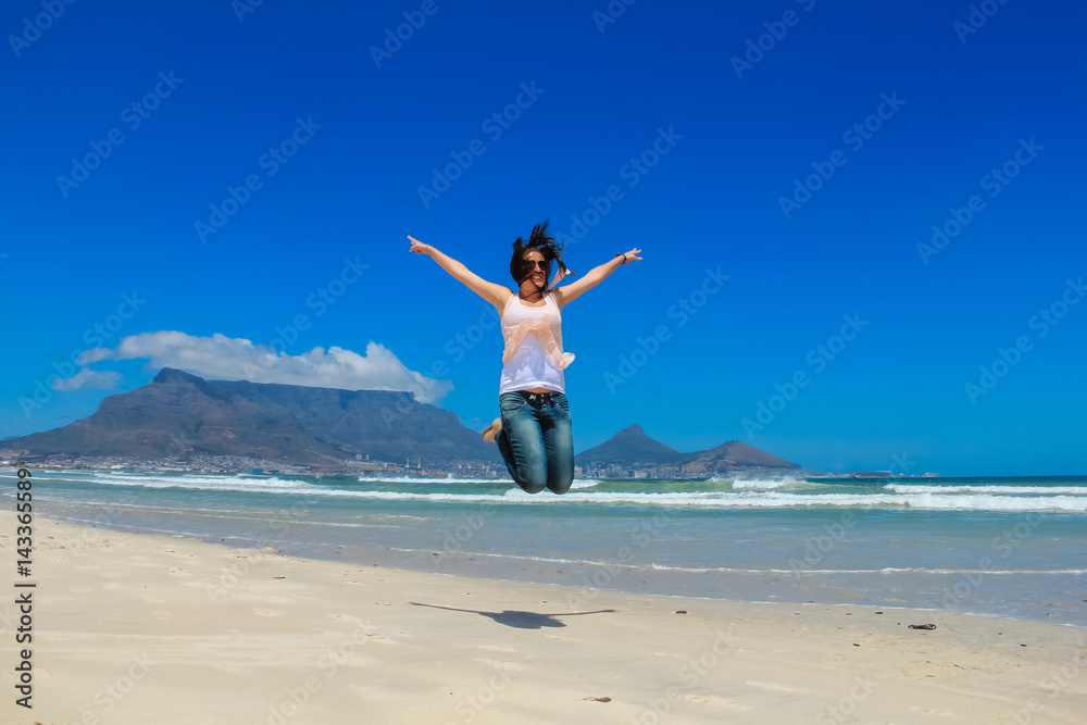Beautiful girl jumping at Milnerton beach, Cape Town. Table Mountain and breaking waves in the background. Concept photo of happiness, freedom and lifestyle. Beautiful girl jumping at Milnerton beach, Cape Town. Table Mountain and breaking waves in the background. Concept photo of happiness, freedom and lifestyle.