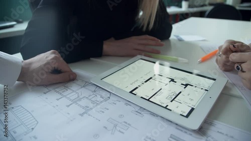 Two women and man discuss architectural project in office using laptop. Three people work in team, now it is discussion. In front of them paper with drawing lay. Also on white plastic table in its