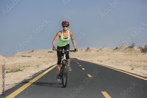 Closeup of beautiful woman wearing a bicycle helmet and sports glasses with tattoos riding a mountain bike on the road with a desert background  © Paul